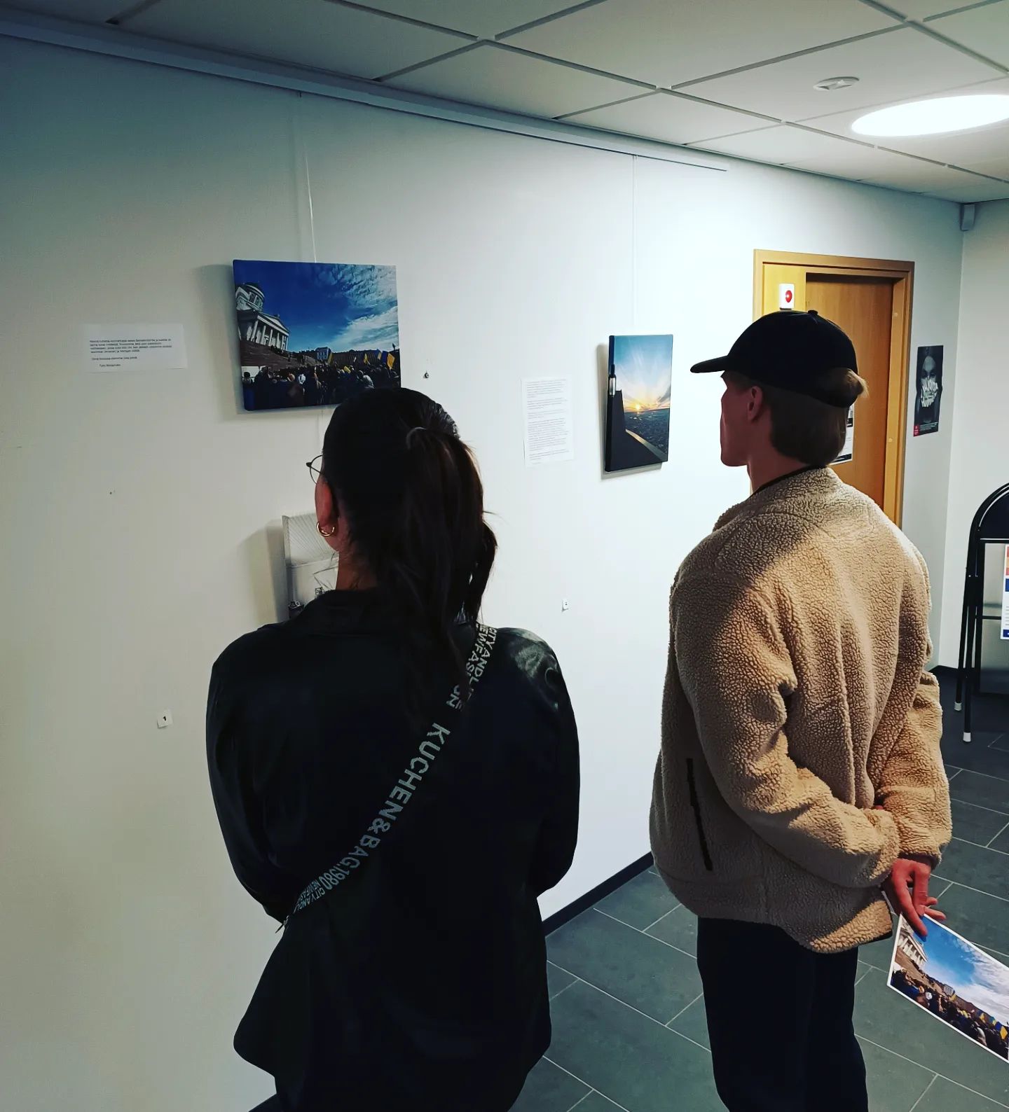 Kaksi henkilöä tarkastelee taideteoksia näyttelyssä, seinällä on valokuvia ja tekstejä.