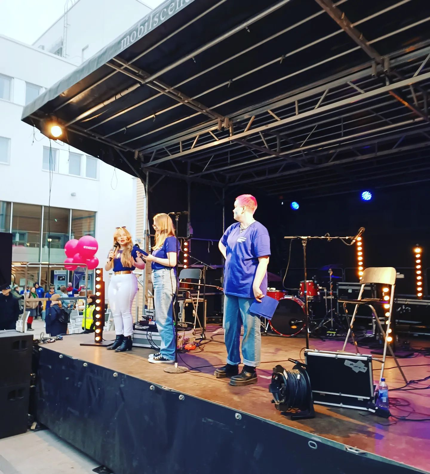 Kolme henkilöä esiintyy lavalla, taustalla näkyy musiikkivälineitä ja yleisöä.