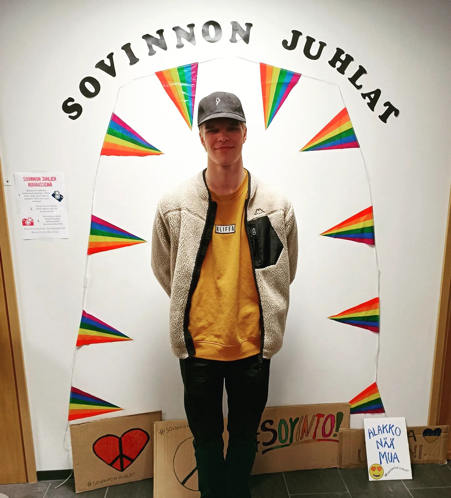Nuori mies seisoo värikkään sateenkaarikoristeen edessä, taustalla teksti "SOVINNON JUHLAT" ja käsintehtyjä kylttejä.
