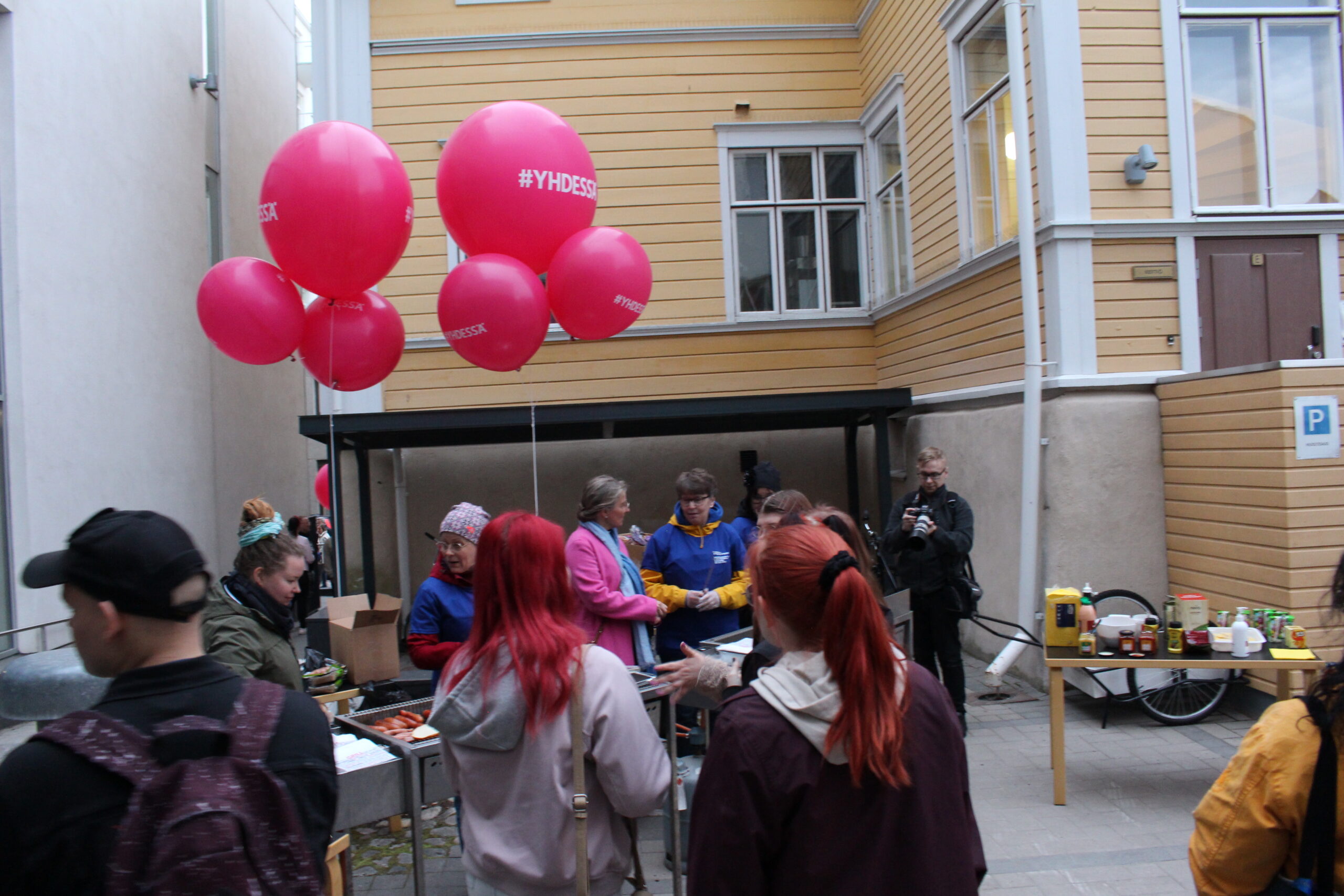 Kuvassa on tapahtuma, jossa osallistujat keskustelevat ja nauttivat grilliruokaa, taustalla näkyy vaaleankeltaisia rakennuksia ja pinkkejä ilmapalloja.