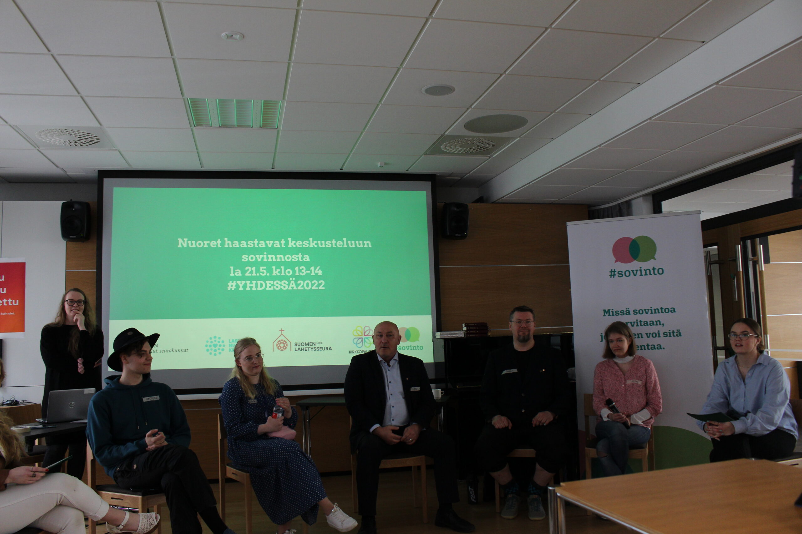 Nuoret keskustelemassa sovinnosta tapahtumassa, taustalla esitys ja mainosbanneri.