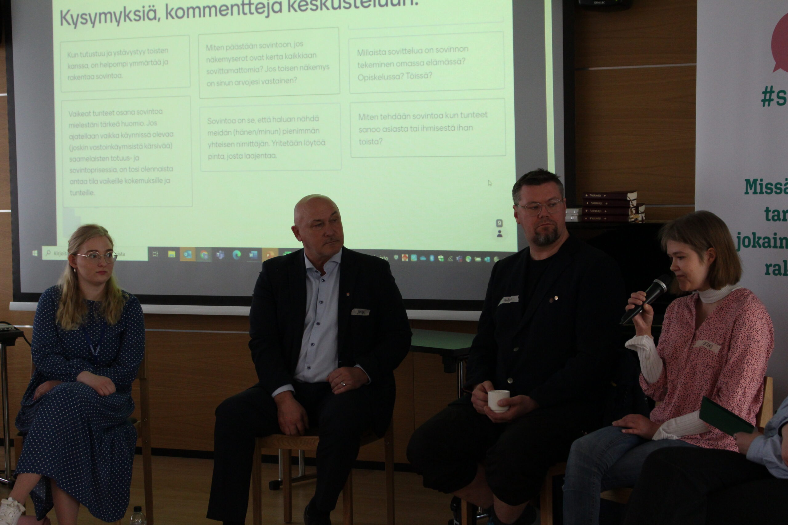 Kuvassa on neljä henkilöä keskustelemassa, taustalla esitys, jossa kysymyksiä ja kommentteja aiheesta sovinto.