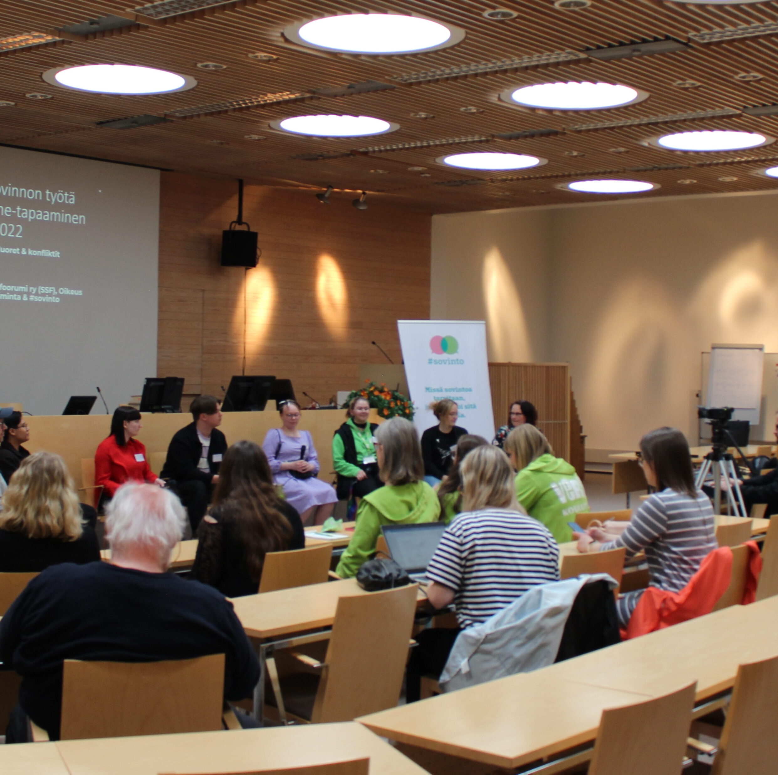 Kuvassa on paneelikeskustelu, jossa useita henkilöitä istuu lavalla ja yleisö seuraa tapahtumaa.