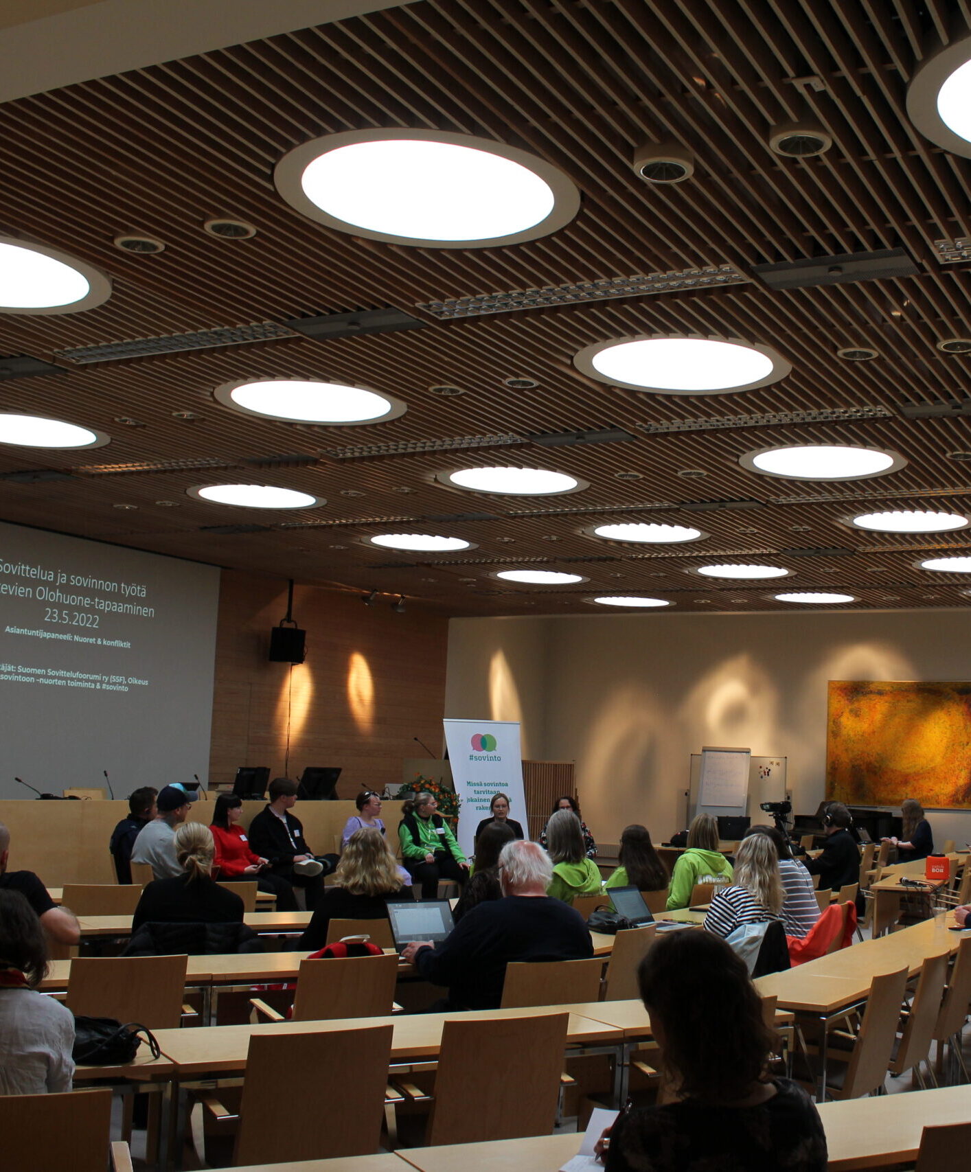 Kuvassa on konferenssitilaisuus, jossa osallistujat istuvat pöydän ympärillä ja keskustelemassa, taustalla näkyy esitysnäyttö.