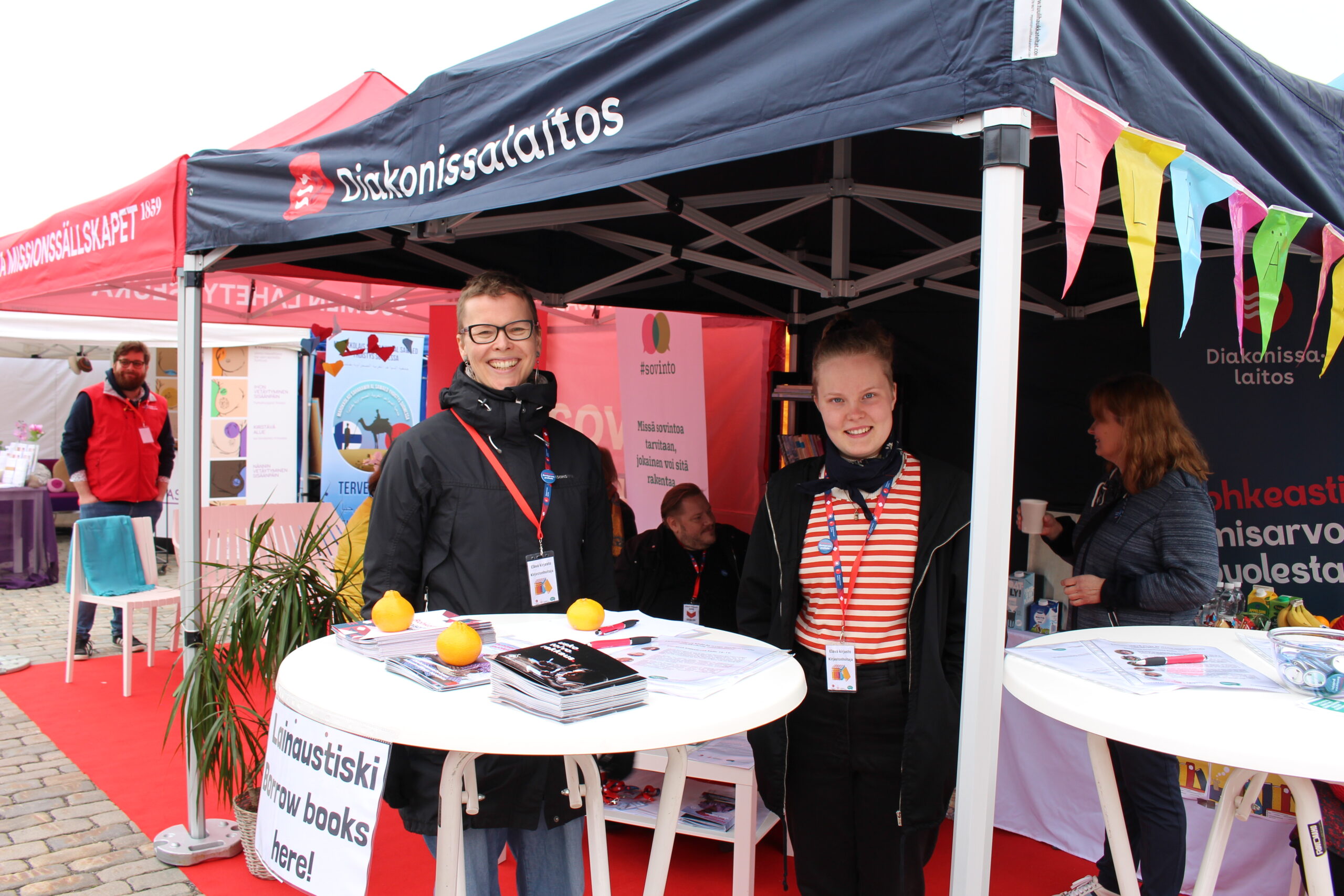 Kuvassa on kaksi nuorta henkilöä hymyilemässä messupisteellä, jossa on esillä esitteitä ja hedelmiä, taustalla näkyy värikkäitä banderolleja ja muita messuosast