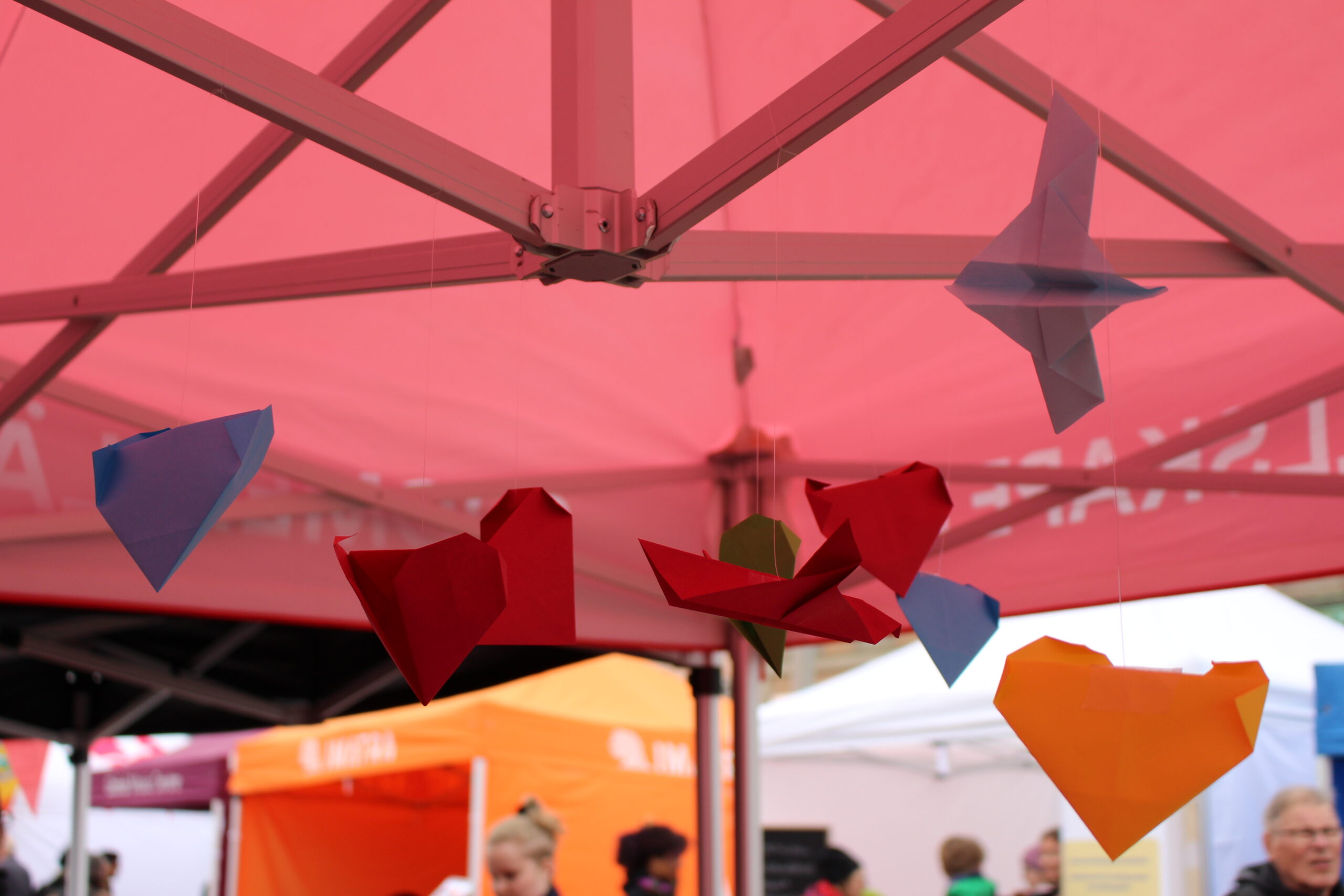 Kuvassa on värikkäitä paperisia sydämiä ja lentokoneita roikkumassa vaaleanpunaisen teltan alta.