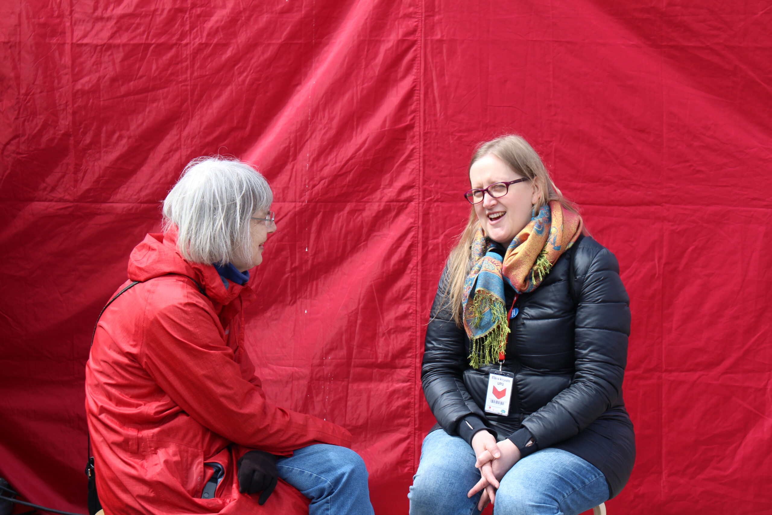 Kaksi naista istuu keskustelemassa punaisen taustan edessä.