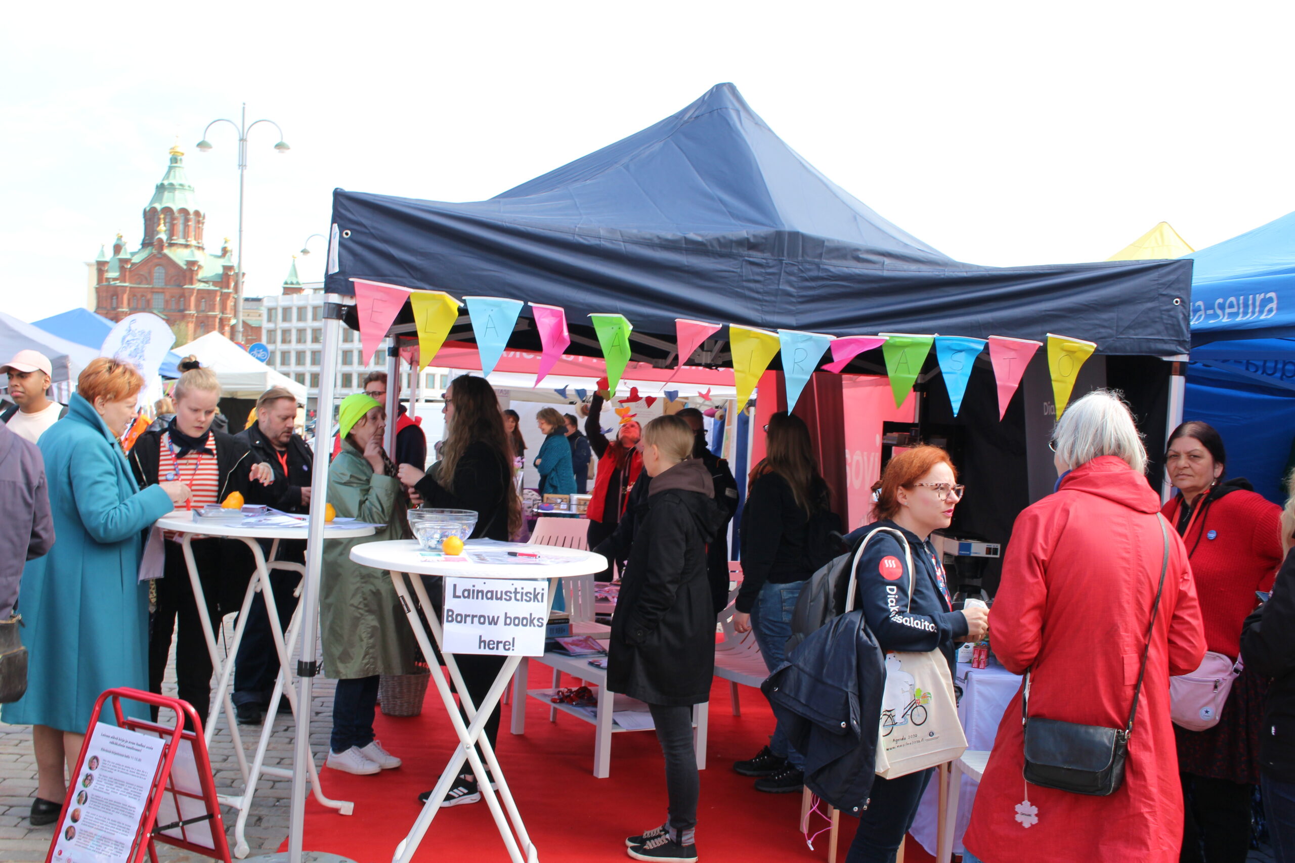 Kuvassa on vilkas tapahtuma, jossa ihmiset keskustelevat ja tutustuvat eri kojujen tarjontaan, taustalla näkyy koristeellisia telttoja ja kaupunkinäkymä.