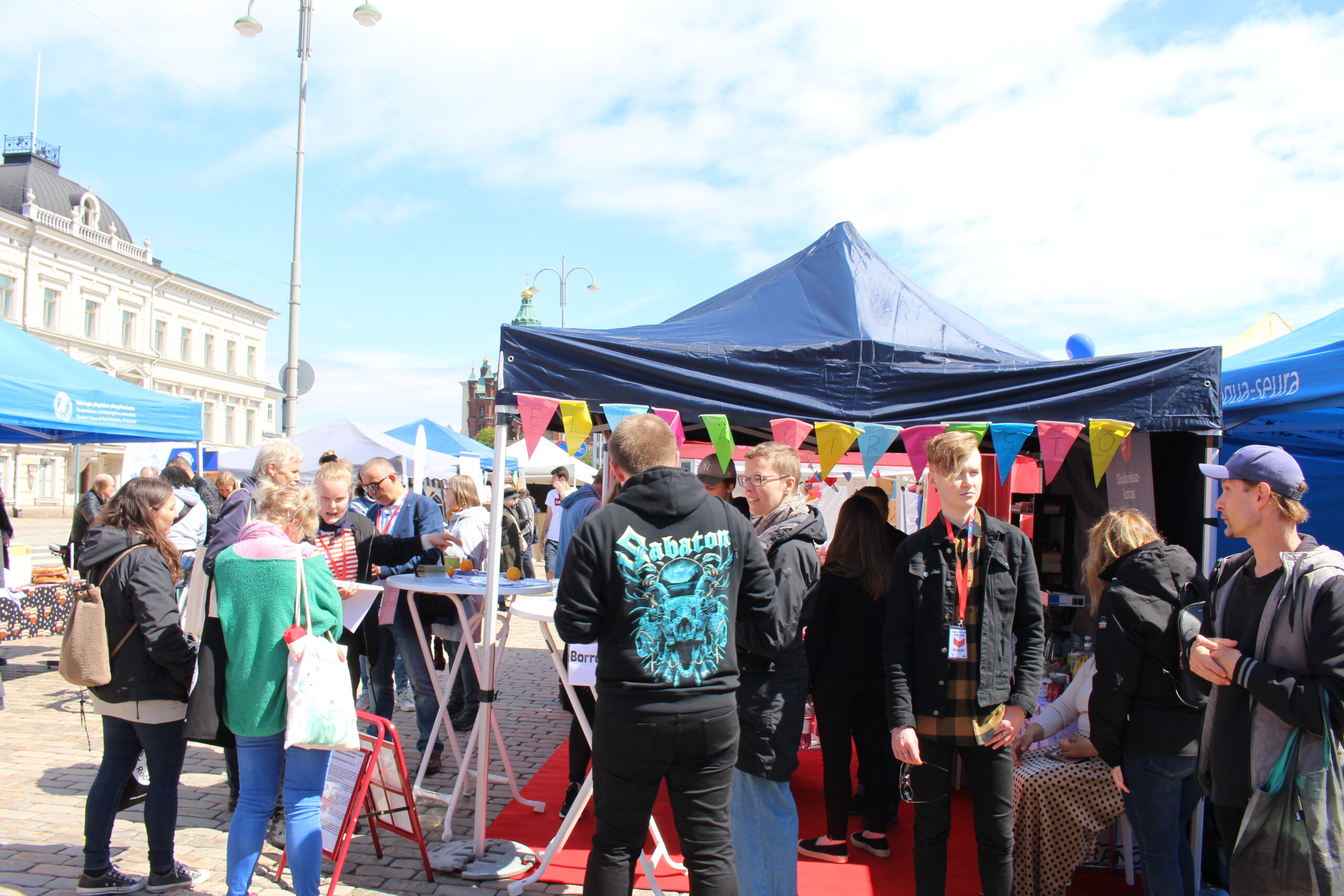 Kuvassa on vilkas tapahtuma, jossa ihmisiä on useita eri kojujen ympärillä ja värikkäitä viirejä koristamassa aluetta.