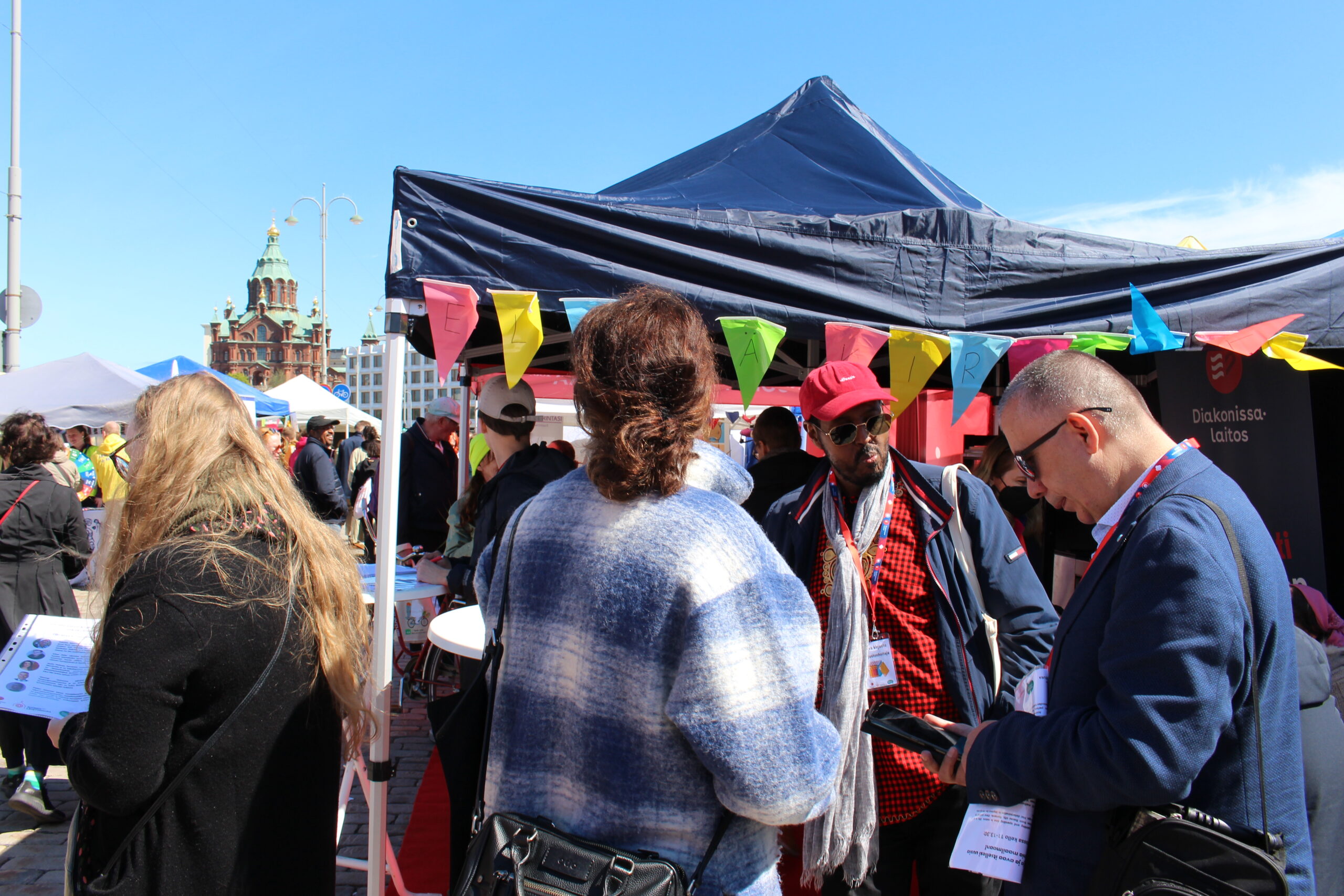 Kuvassa on vilkas tapahtuma, jossa ihmiset keskustelevat ja tutustuvat eri kojujen tarjontaan värikkäiden viirien alla.