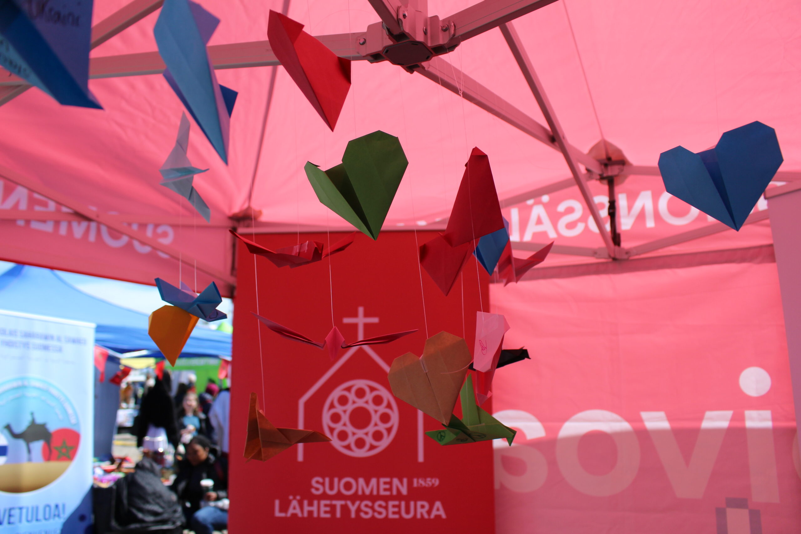 Kuvassa on värikkäitä paperilentoja roikkumassa vaaleanpunaisessa teltassa, jonka taustalla näkyy Suomen Lähetyssäätiön logo.